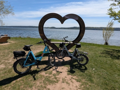 Zwei E‑Bikes stehen auf einer Wiese vor einem großen herzförmigen Rahmen. Dahinter ist ein See mit blauem Himmel und etwas Uferlandschaft zu sehen.